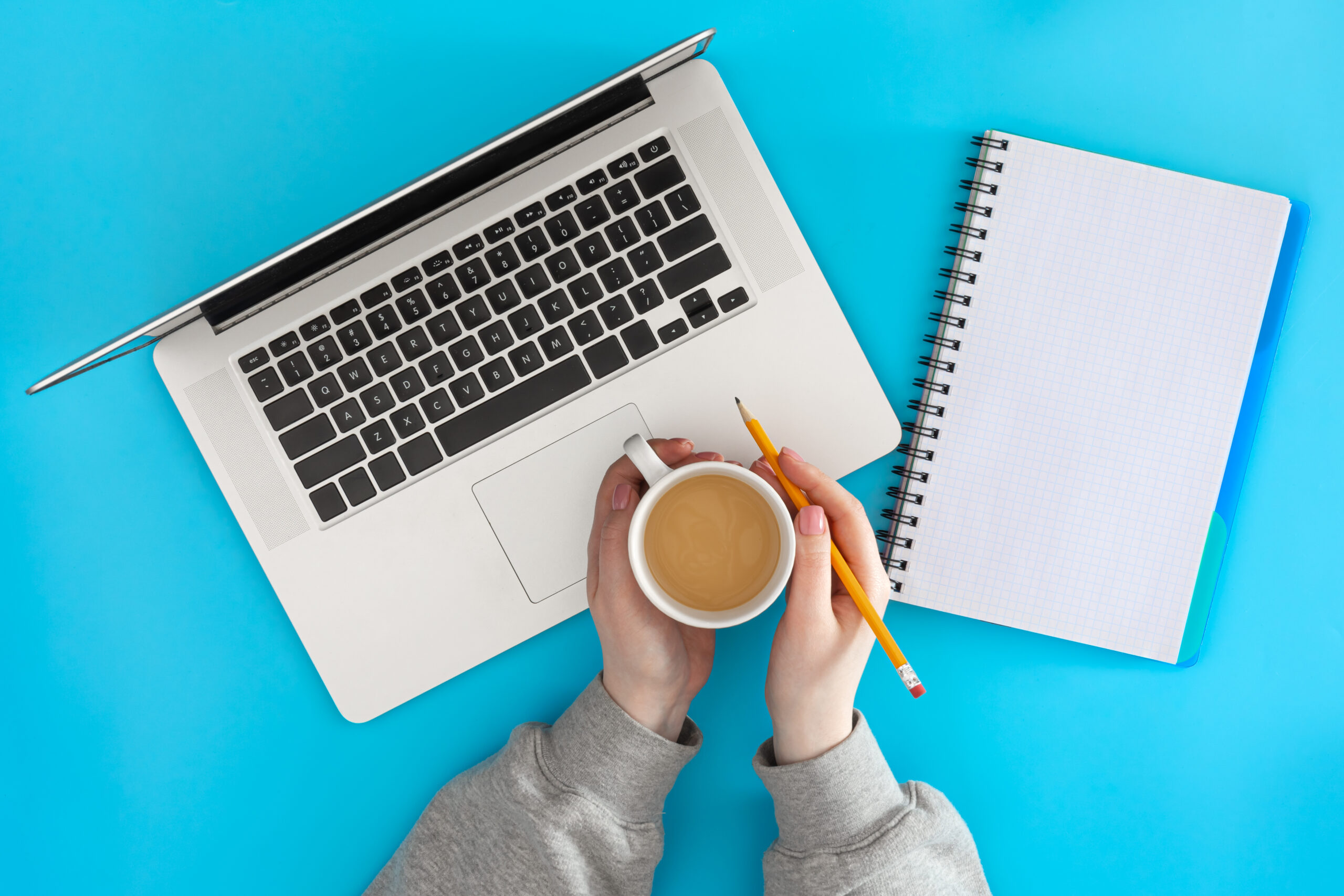 A top-down view of a minimalist freelancer workspace on a vibrant blue background, featuring a laptop, an open spiral notebook, a pencil, and hands holding a cup of coffee