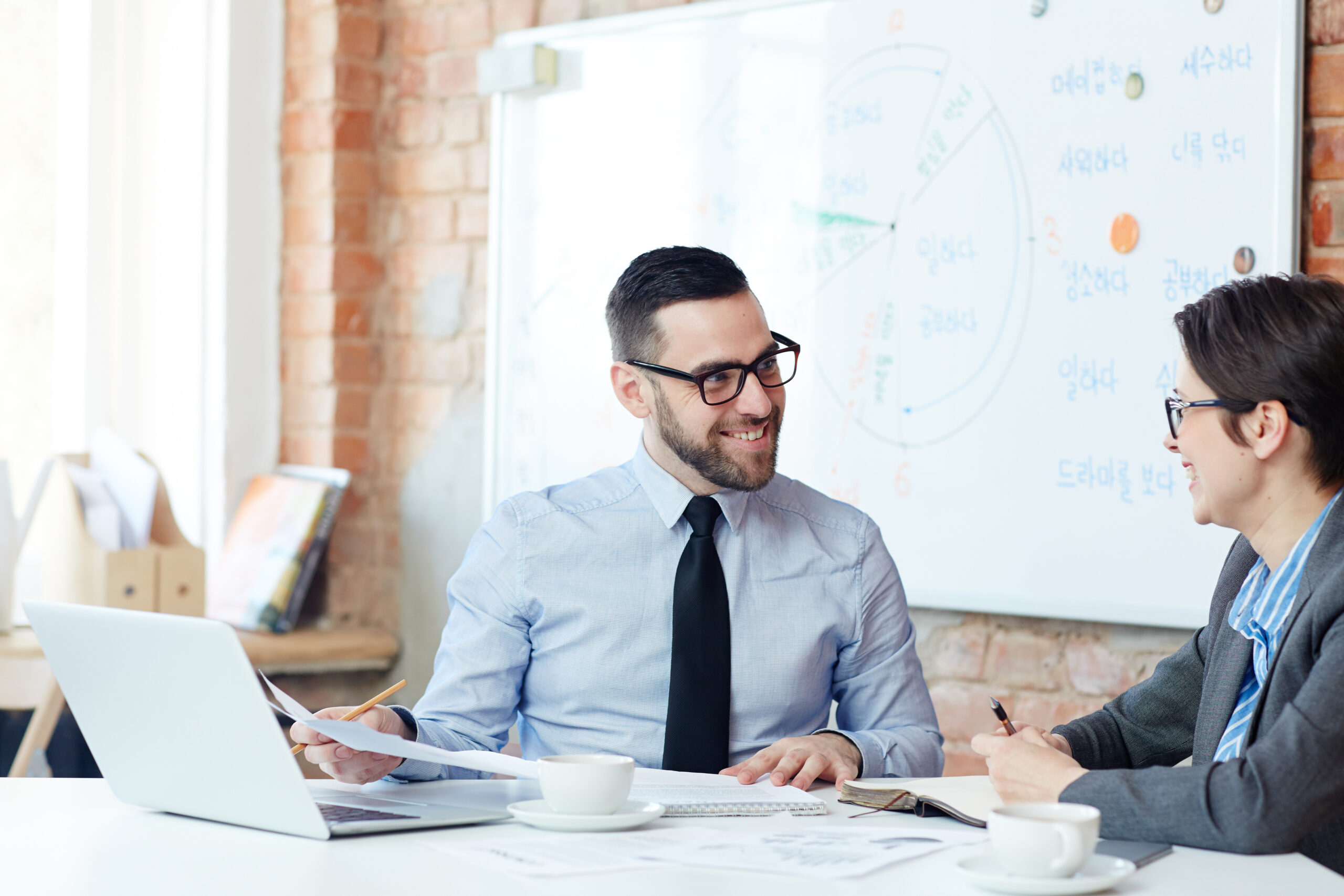 A smiling professional male consultant in a tie and glasses discussing strategy with a client over coffee, with a laptop and a brainstorming whiteboard in the background.